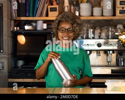 Ritratto di sorridente cameriera afroamericana con capelli afro guardando la macchina fotografica con shaker cocktail in discoteca pub Foto Stock