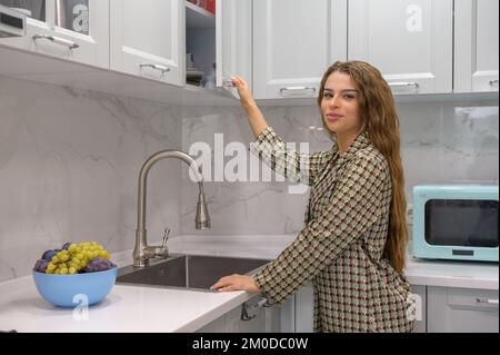 La giovane casalinga carina apre la porta dell'armadio della cucina Foto Stock