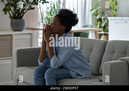 Scoraggiata donna afro-americana penosa guardando in lontananza si siede sul divano nel soggiorno Foto Stock