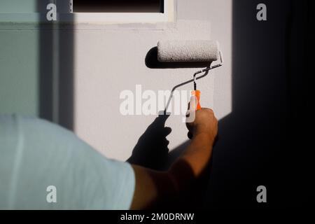 Parete per verniciatura a mano maschio con rullo per verniciatura. Dipingere la stanza delle pareti, ristrutturando con vernice di colore grigio, la mano dell'operaio dipinge la parete con un rullo in gre Foto Stock