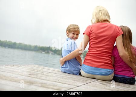 Tempo di qualità con i miei figli. Un'immagine posteriore di una madre con suo figlio e sua figlia seduti accanto ad un fiume con spazio di copyright. Foto Stock