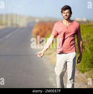 Indovino che Ill debba hitchhike. Un giovane che cerca di agganciare un giro mentre cammina lungo una strada deserta. Foto Stock