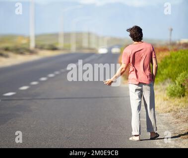 Indovino che Ill debba hitchhike. Un giovane che cerca di agganciare un giro mentre cammina lungo una strada deserta. Foto Stock