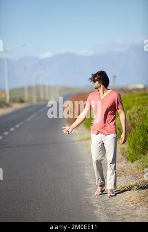 Indovino che Ill debba hitchhike. Un giovane che cerca di agganciare un giro mentre cammina lungo una strada deserta. Foto Stock