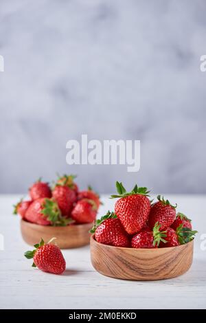 Fragole mature in due ciotole di legno su un tavolo di legno bianco su sfondo grigio. Verticale, spazio di copia, primo piano. Foto Stock
