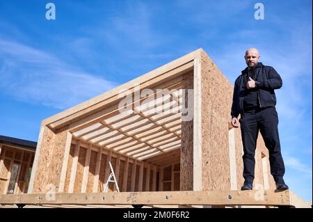 Sviluppatore costruzione di case con struttura in legno in stile scandinavo barnhouse. Uomo calvo in piedi in cantiere, ispezionando la qualità del lavoro, mostrando pollici in su nelle giornate di sole con cielo blu sullo sfondo. Foto Stock