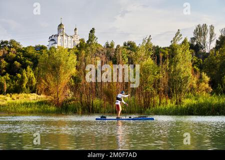 Uomo a stand up paddle tavole SUP nel lago di montagna Sairan nella città di Almaty vicino alla Chiesa ortodossa in Kazakistan. Foto Stock