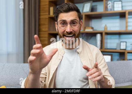 Insegnante maturo che insegna in remoto, uomo con occhiali e barba guardando web camera e sorridendo parlando con i colleghi e l'insegnamento, uomo d'affari a casa seduto sul divano in soggiorno. Foto Stock