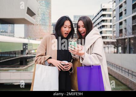 Due giovani donne felice multietnico sorpreso circa le vendite e offre cercando un telefono cellulare facendo shopping online. Consumatori Signore amici che acquistano vestiti con lo smartphone. Concetto di e-commerce. Foto di alta qualità Foto Stock