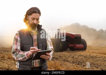 Un agricoltore con tablet digitale controlla un trattore autonomo in un'azienda agricola intelligente Foto Stock