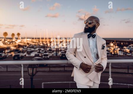 Un uomo nero bearded elegante posa all'ora d'oro; l'uomo africano è vestito in un vestito beige con cravatta blu dell'arco e punti bianchi della polka e sunglasse Foto Stock