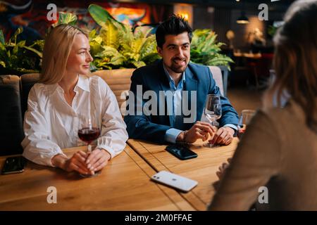 Elegante barbuto uomo in tuta e due belle donne bionde seduto ristorante parlare ridendo fare toast divertirsi, celebrare il compleanno. Foto Stock