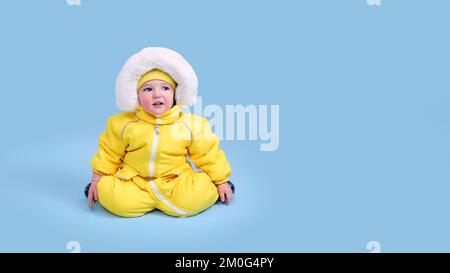 Bambino felice in abiti invernali muta di neve su sfondo blu studio. Un bambino in una tuta gialla calda con cappuccio. Bambino di un anno e cinque mesi Foto Stock