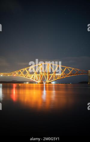 Splendido scenario del famoso Forth Rail Bridge illuminato da luci notturne riflesse nell'acqua in Scozia, Regno Unito, sotto il cielo stellato Foto Stock