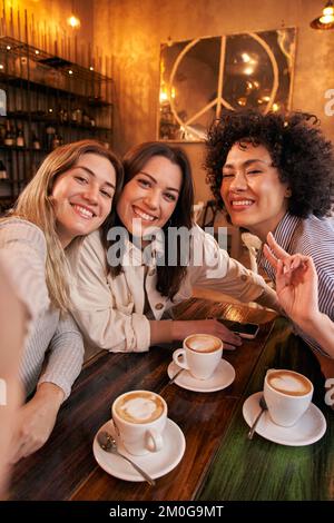 Foto verticale di tre amici femminili prendere selfie mentre si diverte in caffetteria. Felice sorridente ragazze guardando la macchina fotografica Foto Stock