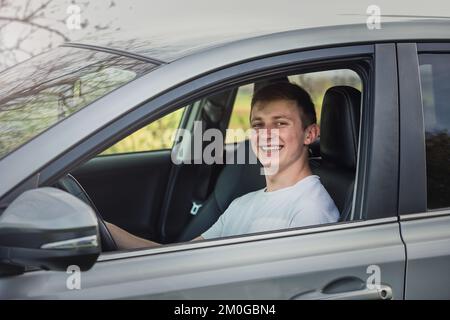 Giovane uomo gioioso che guida sicuro la sua nuova auto, cercando allegro alla macchina fotografica. Il conducente giovane soddisfatto che si gode la guida, tiene le mani sul volante t Foto Stock