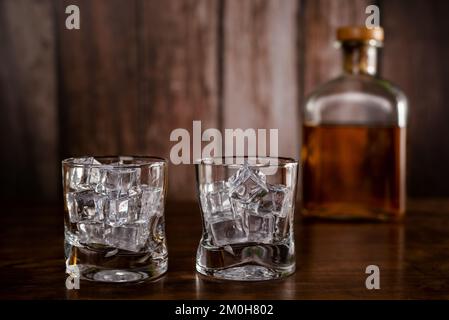 Due bicchieri di whiskey vuoti con cubetti di ghiaccio, con una bottiglia con whiskey su fondo sfocato. Foto Stock