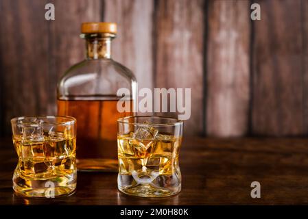 Due bicchieri di whiskey con cubetti di ghiaccio, con una bottiglia con whiskey sullo sfondo fuori fuoco. Foto Stock