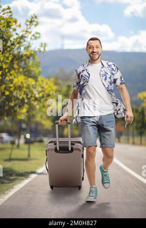 Ritratto completo di un uomo sorridente che cammina e tira una valigia in un parco Foto Stock