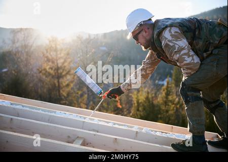 Costruttore maschile che fa isolamento termico sul tetto di casa con telaio in legno la sera. Uomo lavoratore spruzzando schiuma di poliuretano sul tetto del futuro cottage. Concetto di costruzione e isolamento. Foto Stock