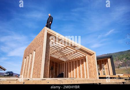Costruttore maschile costruzione casa con telaio in legno. Uomo calvo in piedi sul cantiere, che ispeziona la qualità del lavoro nelle giornate di sole con il cielo blu sullo sfondo. Foto Stock