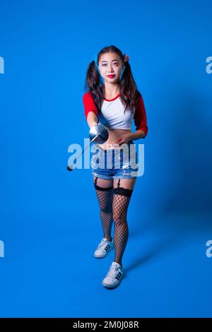 Giovane donna in Harlequin Costume Holding Fencing foil contro uno sfondo blu solido Foto Stock