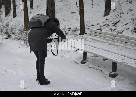 giovane ragazza fotografa con una borsa grigia scatta foto di una panca di legno innevata nel parco. Foto Stock