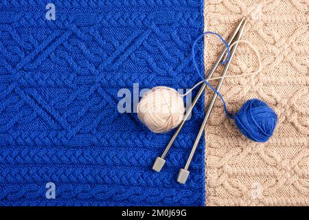 Due palle di filo e aghi per maglieria giacciono su un tessuto lavorato a maglia blu e beige. Il concetto di passare del tempo a casa e lavorare a maglia abiti caldi per wi Foto Stock