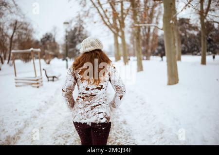 Vista da dietro donna di mezza età in cappello marrone e sciarpa in abiti nevosi e cappotto di pelle di pecora fuori in città in inverno. Foto Stock