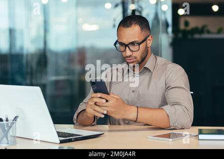 Scioccato e sorpreso giovane afro-americano seduto in ufficio al tavolo e tenendo il telefono in mano, ha ricevuto un messaggio, leggere le notizie. Foto Stock