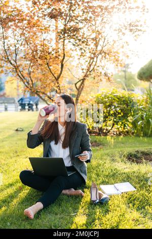 Bella donna che si rilassa bevendo una tazza di caffè dopo il lavoro duro e utilizzando il computer portatile mentre si siede sull'erba verde nel parco. Tecnologia e libertà Foto Stock