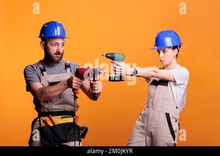 Team di operai edili che tengono pistole di perforazione in studio, posando con utensili elettrici a vite in background. Uomo e donna che usano il trapano elettrico e indossano tute con elmetto, cintura portattrezzi. Foto Stock