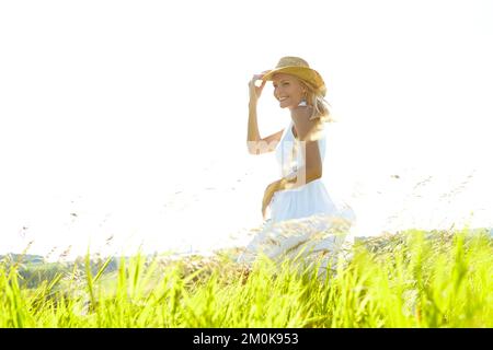 Sia sempre una ragazza di paese. Una bella giovane donna bionda in piedi in un prato che indossa un cappello da sole in una calda giornata estiva. Foto Stock