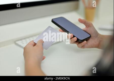 Immagine ravvicinata, Una mano femminile che tiene uno smartphone e una carta di credito sopra la scrivania. Foto Stock