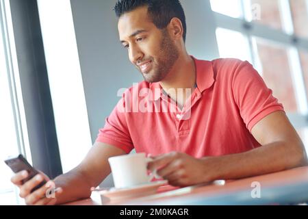 Risposta a un messaggio di testo. Un giovane che legge un SMS mentre si gusta una tazza di caffè. Foto Stock