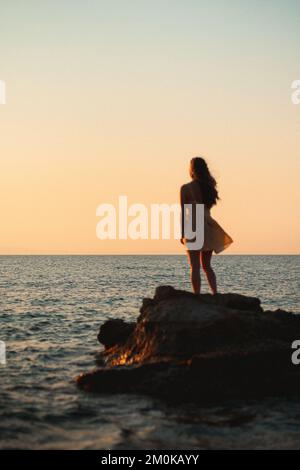 Una giovane donna che guarda l'alba sull'oceano in un bel vestito giallo guardando Foto Stock