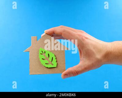 Mano che tiene una casa di cartone con foglia verde all'interno su sfondo blu, immagine concettuale di efficienza energetica Foto Stock
