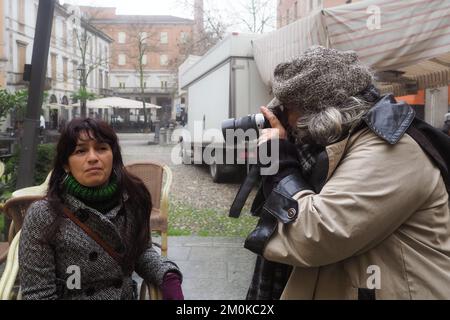 giovane adulta ispanica donna da sola all'aperto in un bar in inverno indossando guanti di lana verde polo v collo, giacca grigia pesante fotografata da femmina senior pho Foto Stock