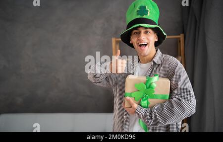 Uomo felice in leprechaun cappello che tiene la scatola regalo del giorno del St.Patrick che guarda la macchina fotografica che mostra i pollici in su come nel paese. Consegna rapida il giorno di San Patrizio Foto Stock