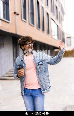 Vita in su ritratto dell'uomo indiano è in piedi con caffè per andare e salutare qualcuno con la mano Foto Stock