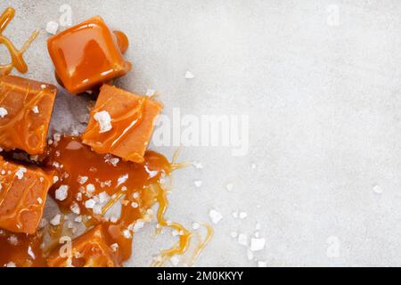 Caramello salato, una sensazione di gusto. Sale grossolano e blocchi di caramello ricoperti in una salsa di caramello salato appiccicoso su grigio Foto Stock