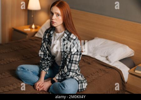 Ritratto di triste giovane donna rossa seduta a letto da sola guardando la macchina fotografica, sentendosi sola a casa. Infelice zenzero femmina ha problemi psicologici. Foto Stock