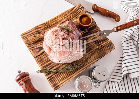 Un grosso pezzo di prosciutto di maiale si trova su un tagliere di carne di legno testurizzato con ingredienti per la cottura. coltello, forchetta per carne, spezie Foto Stock
