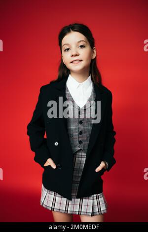 ragazza positiva in giubbotto a scacchi che tiene le mani in tasche di blazer nero isolato su rosso Foto Stock