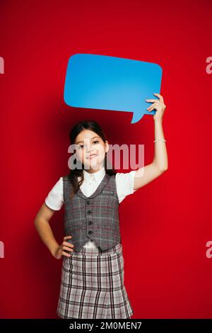 ragazza pretenuta in giubbotto a plaid in piedi con la mano sull'anca e tenendo la bolla di linguaggio blu in mano sollevata isolato sul rosso Foto Stock