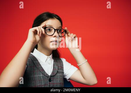 ragazza in giubbotto a plaid guardando lontano e regolando gli occhiali isolati sul rosso Foto Stock