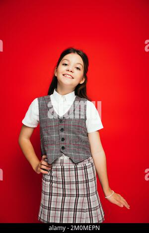 allegra ragazza brunetta in gilet a plaid e gonna in posa con mano sul fianco isolato sul rosso, banner Foto Stock