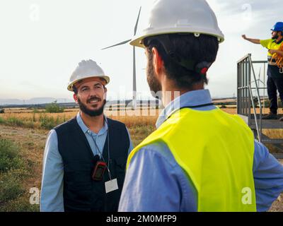 Due ingegneri maschi con caschi protettivi che convergono in un campo di mulini a vento. Energie rinnovabili Foto Stock