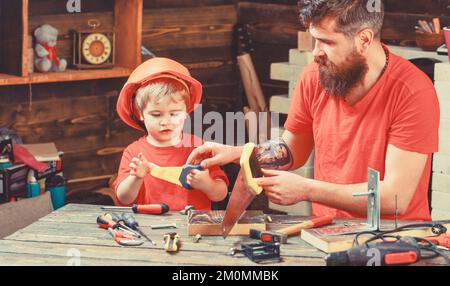 Concetto di paternità. Ragazzo, bambino occupato nel casco protettivo imparare ad usare la sega a mano con papà. Padre, genitore con barba che insegna al piccolo figlio a segare Foto Stock