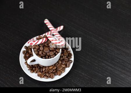 Caramelle natalizie e chicchi di caffè in tazza bianca su tavola di legno scuro. Caffè arrosto scuro, bevanda naturale per le vacanze invernali Foto Stock